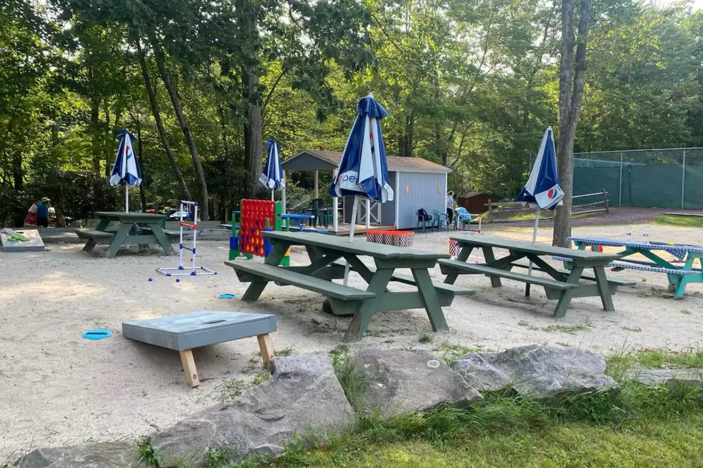 Outdoor sand recreational area featuring green picnic tables with folded umbrellas, cornhole boards, and yard games surrounded by trees.