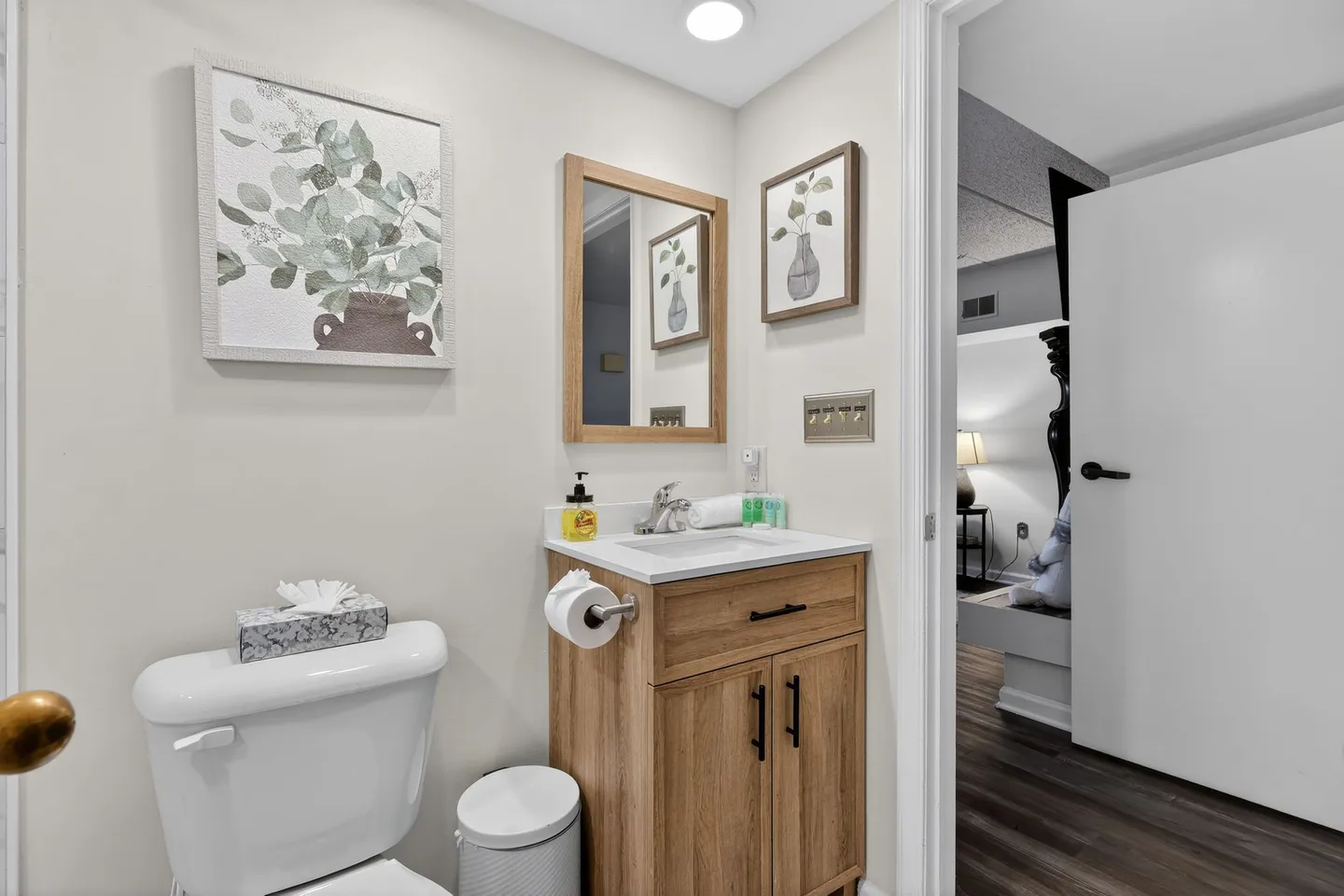 Bathroom vanity with wooden cabinet, white countertop, mirror, and toilet beside an open doorway to a bedroom.