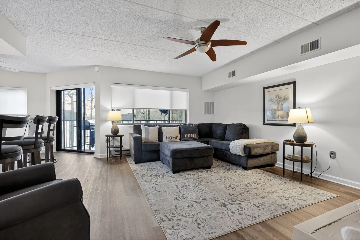 Living room with a dark sectional sofa, ottoman, area rug, wood-look flooring, ceiling fan, and sliding glass door.