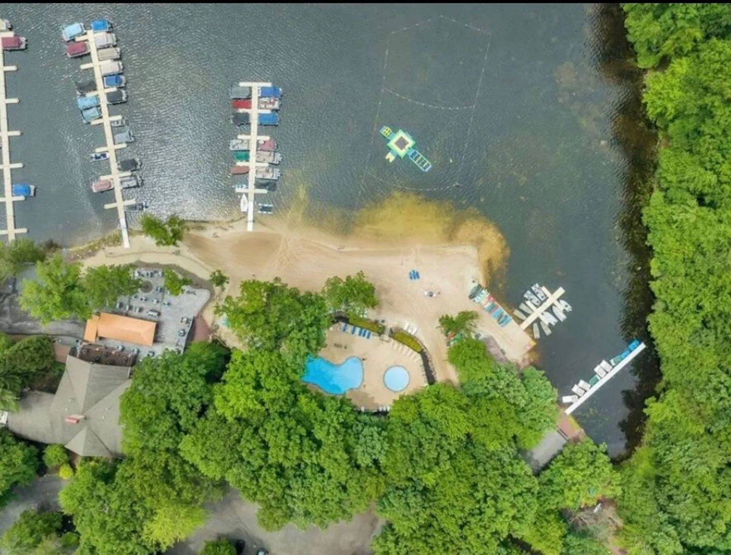 Aerial view of a lakefront resort amenity area featuring swimming pools, a sandy beach, boat docks, and surrounding trees.