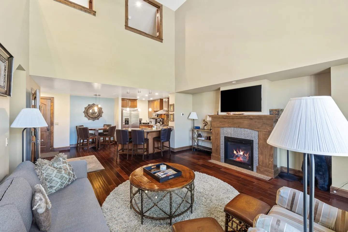 Open living area with vaulted ceilings, a fireplace with a mounted TV above, hardwood flooring, and a view into the kitchen and dining space.