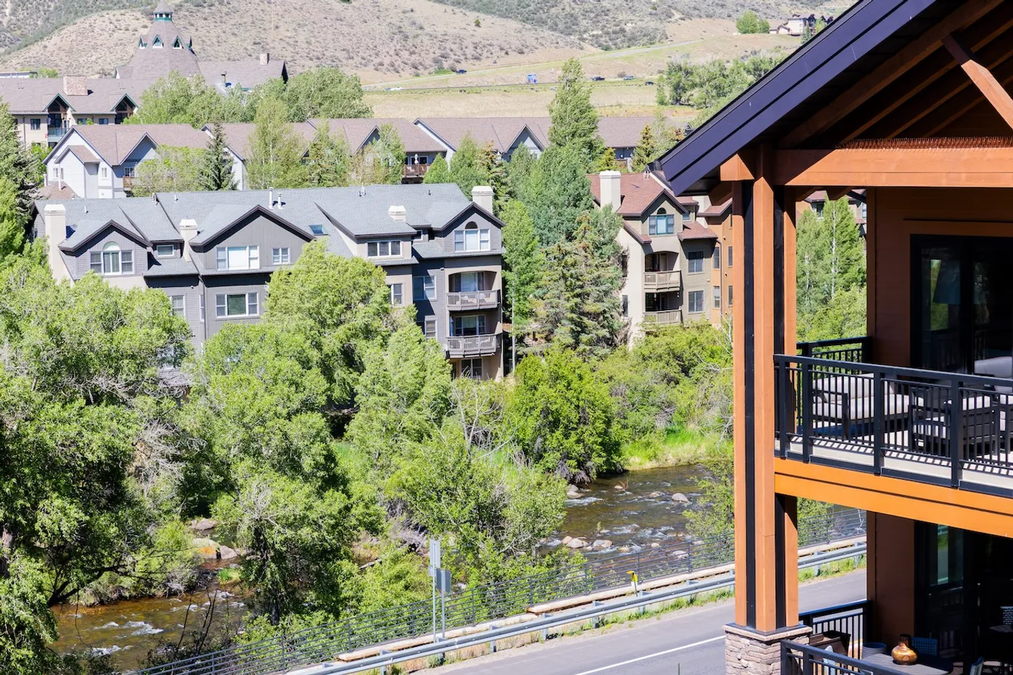 Elevated view from a covered wooden balcony overlooking a flowing river, lush green trees, and nearby multi-story condo buildings.
