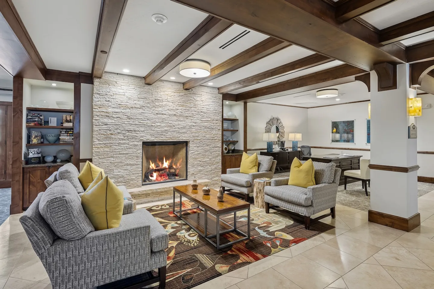 Seating area with armchairs around a coffee table facing a lit fireplace set in a stacked-stone wall beneath wood-beamed ceilings.