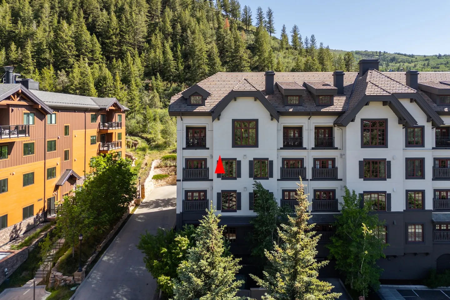 Exterior view of a multi-story lodge-style condominium building backed by a forested hillside, with a red pointer indicating a specific unit window.