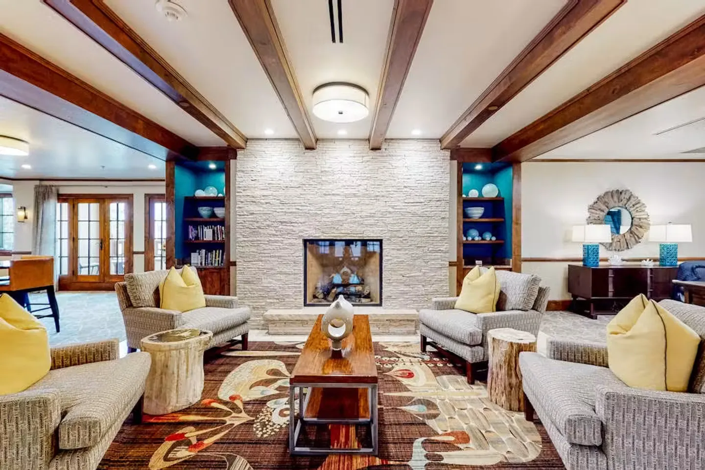Seating area centered around a stone-faced fireplace with exposed wood ceiling beams and patterned rug.