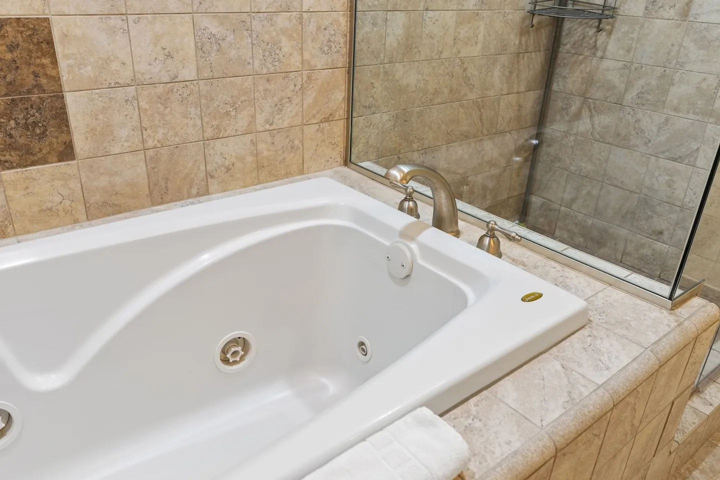 Close-up view of a white drop-in jetted soaking tub surrounded by beige tile next to a glass-enclosed shower.