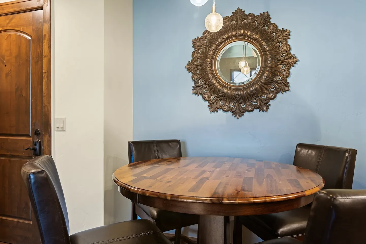 Dining area featuring a round wooden table, dark leather chairs, and an ornate carved mirror on a light blue accent wall.