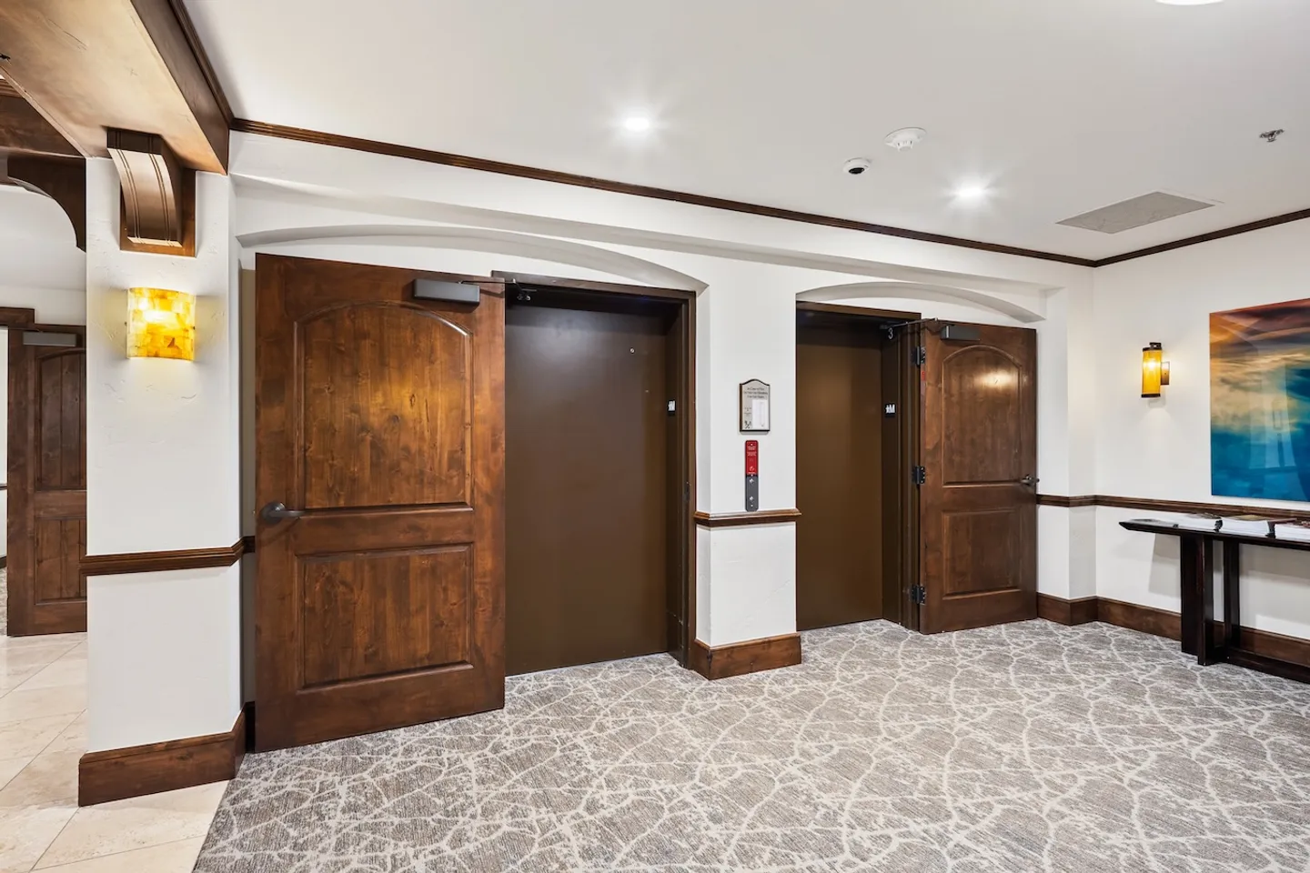 Elevator lobby featuring two elevators framed by arched wooden doors and white walls with wood trim over patterned carpeting.