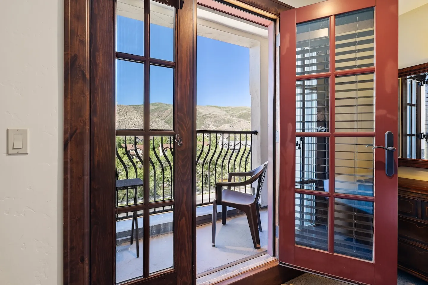 Open French doors looking out to a small private balcony with chairs and views of sunny hills and blue sky.