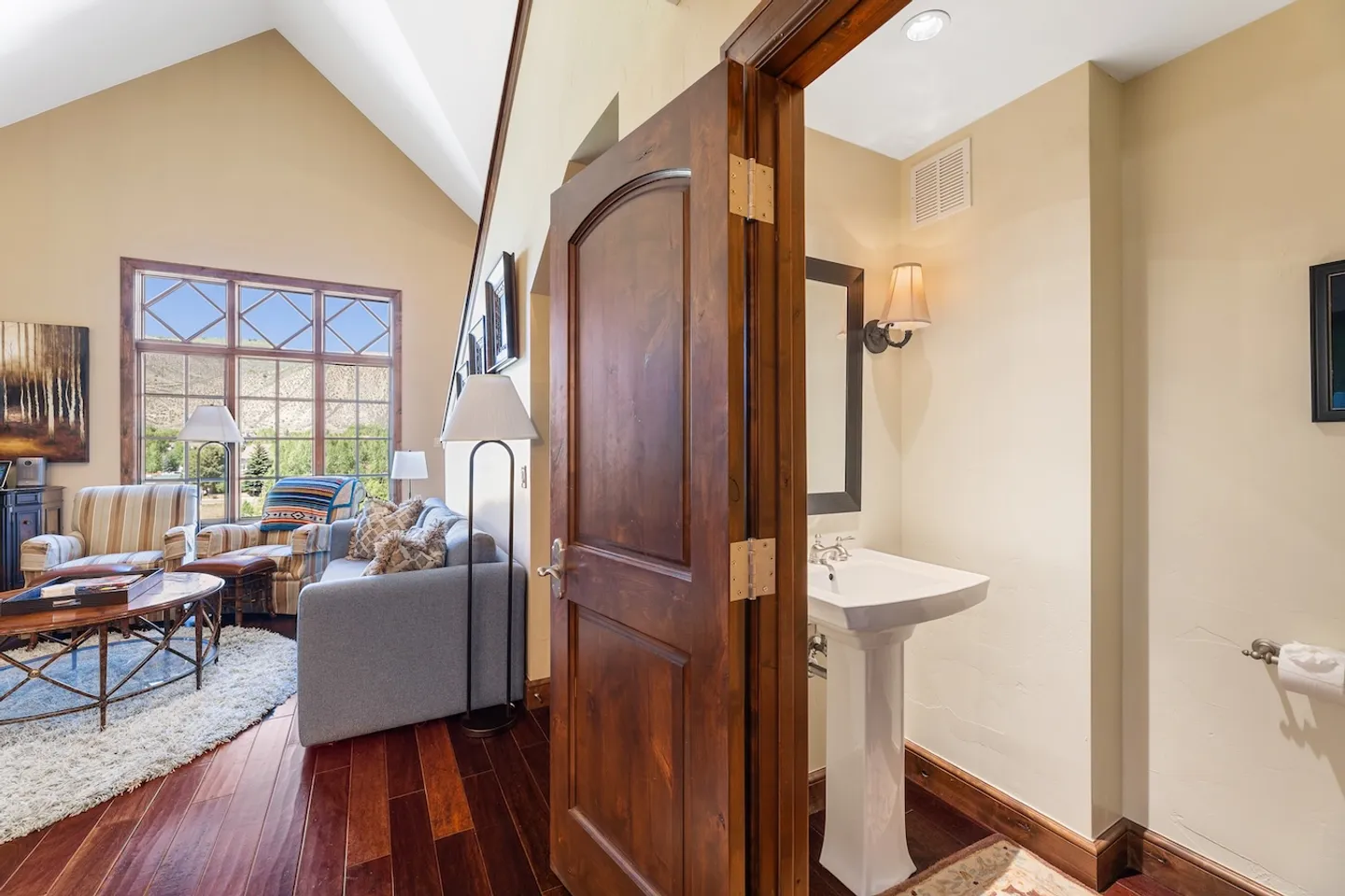 Open wooden door showing a powder room with pedestal sink, leading into a living room with vaulted ceiling.