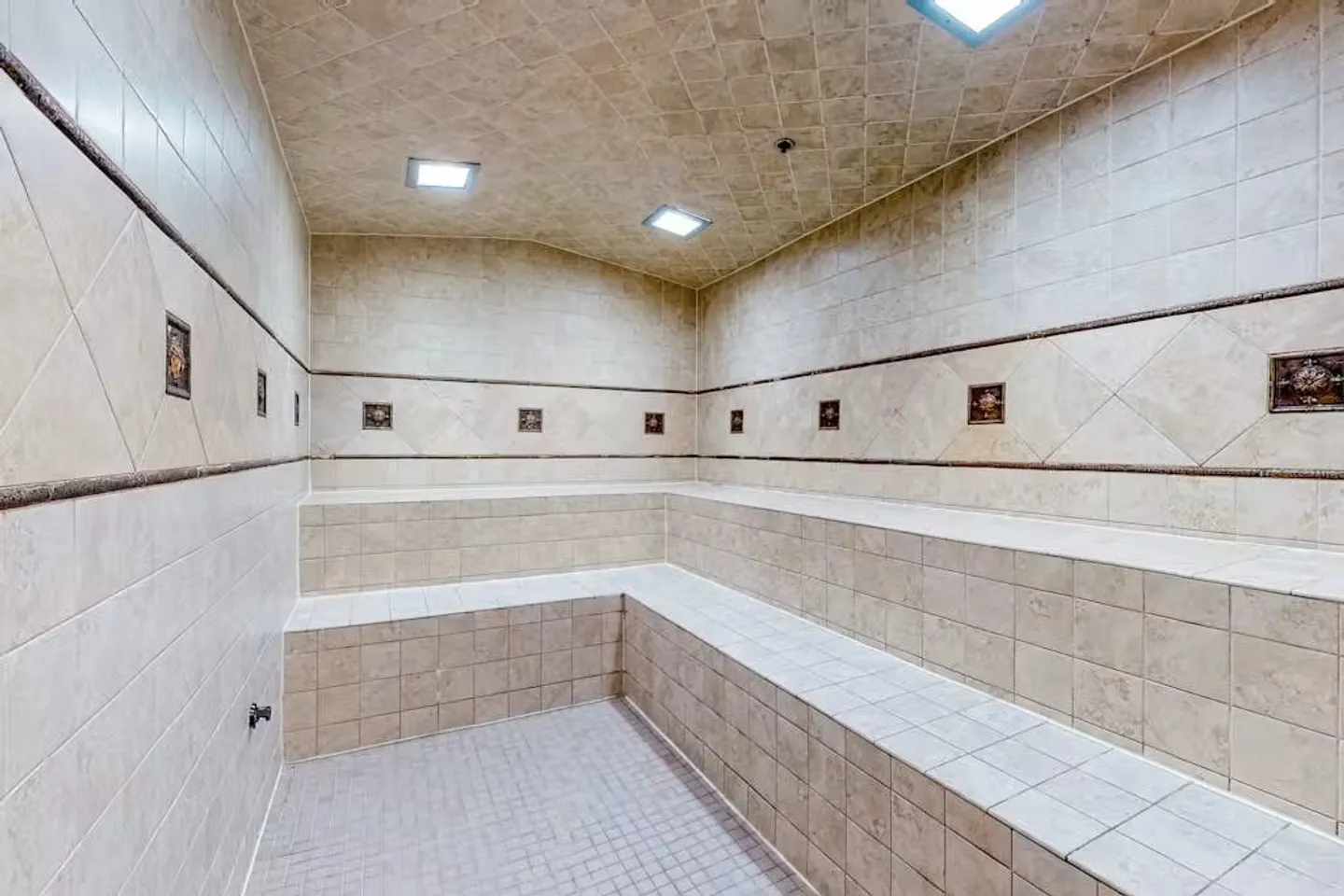 Fully tiled indoor steam room or sauna featuring built-in stepped bench seating and square recessed ceiling lights.