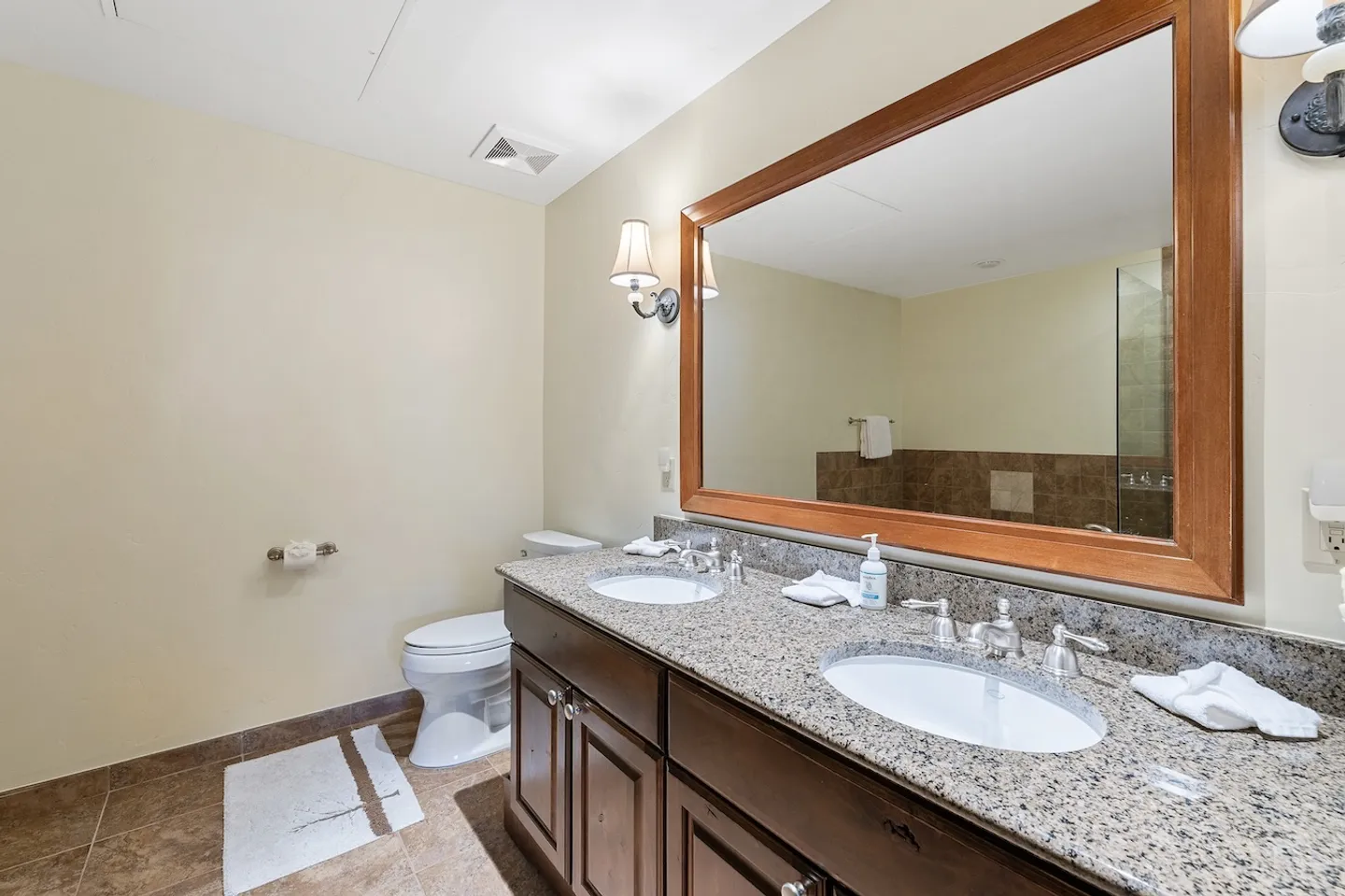 Bathroom with a double-sink dark wood vanity, speckled stone countertop, large wood-framed mirror, and a toilet.