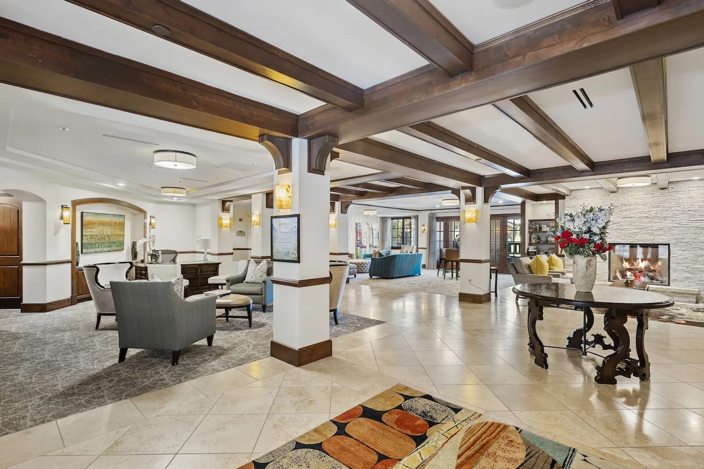 Spacious lobby or common area with dark wood exposed ceiling beams, tile flooring, supporting pillars, and multiple seating arrangements including armchairs and a fireplace.
