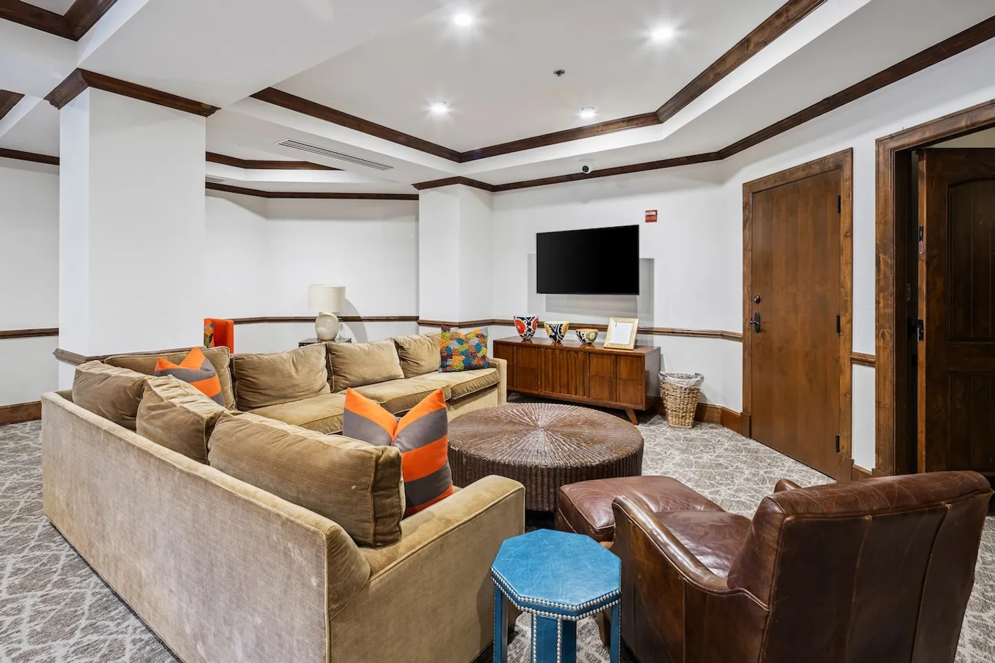 Lounge area featuring a large sectional sofa, round woven coffee table, leather armchair with ottoman, and a wall-mounted TV.