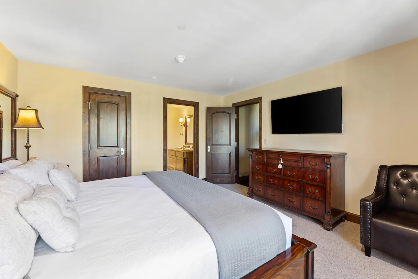 Bedroom featuring a made bed with white and gray bedding, a dark wood dresser beneath a wall-mounted TV, a leather armchair, and doors leading to an ensuite bathroom and closet.
