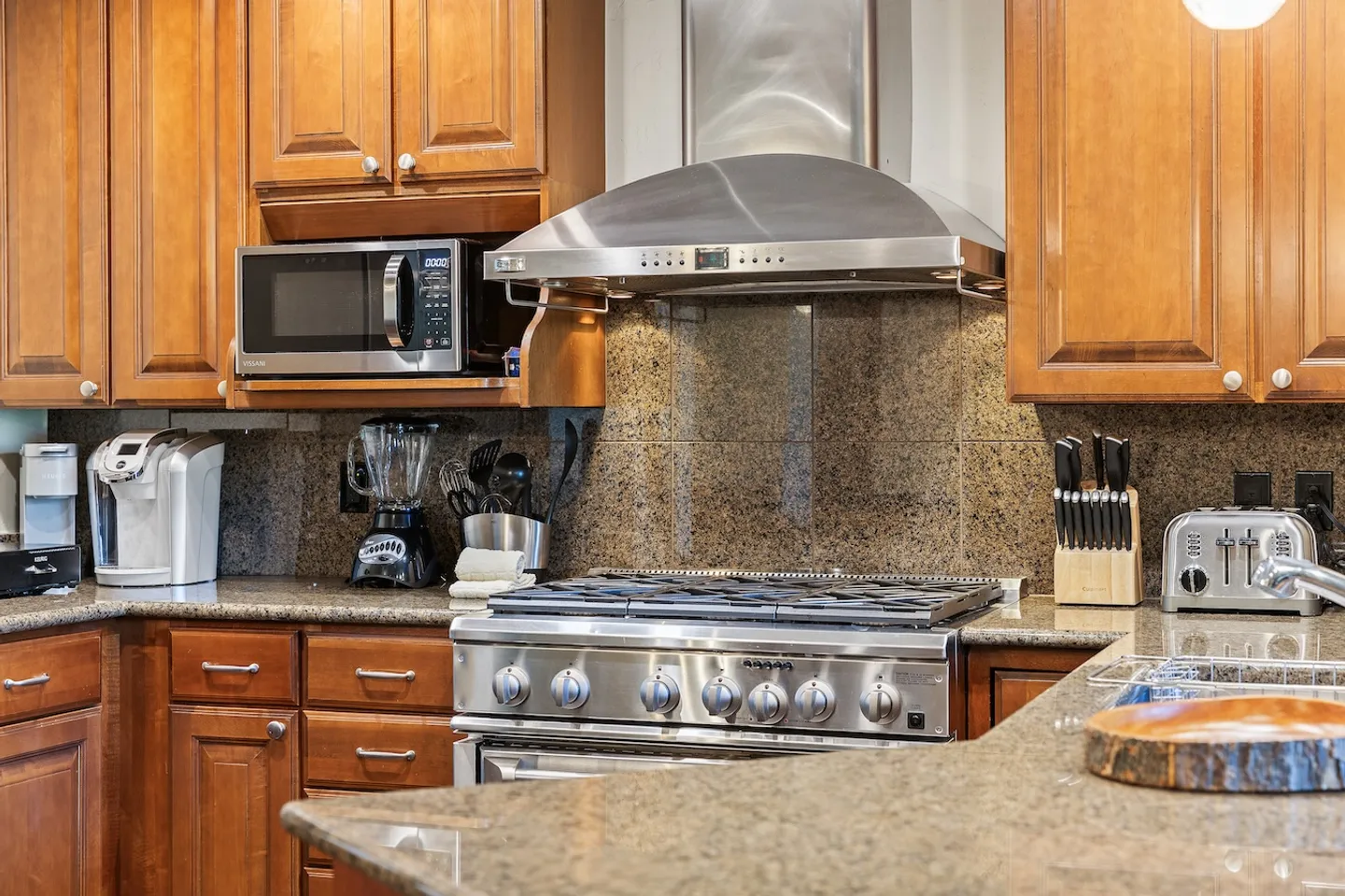 Close-up view of a kitchen featuring wooden cabinetry, speckled granite countertops, a stainless steel range with hood, and small countertop appliances.