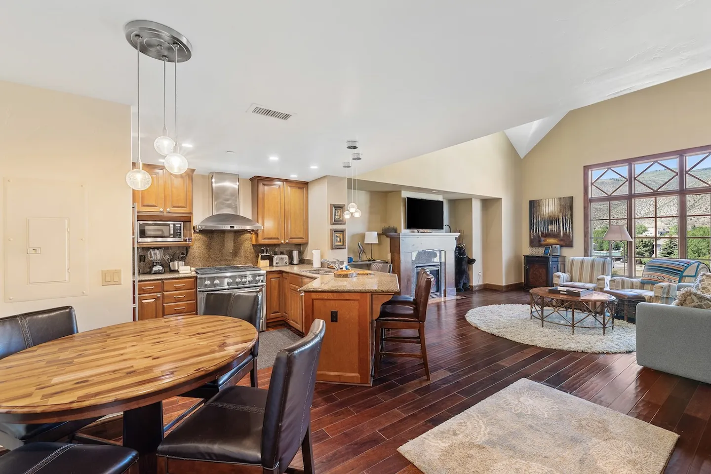 Open-concept living space with a dining table in the foreground, wood-cabinet kitchen with island, and living room with fireplace.