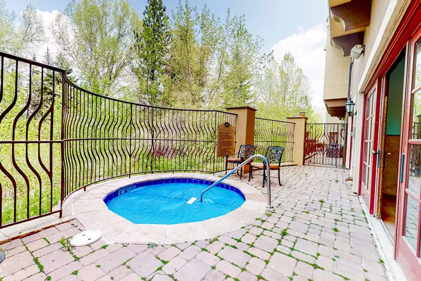 Outdoor paved patio featuring a small circular hot tub or plunge pool with a blue cover, metal railing, and chairs beside a building.