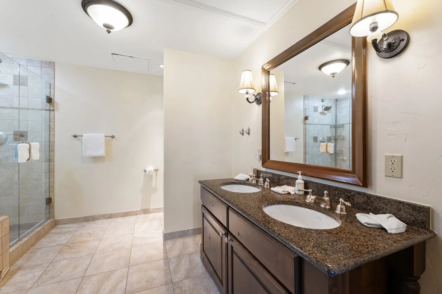 Bathroom with a dark wood double-sink vanity, speckled countertop, framed mirror, tiled floor, and a glass-enclosed shower.