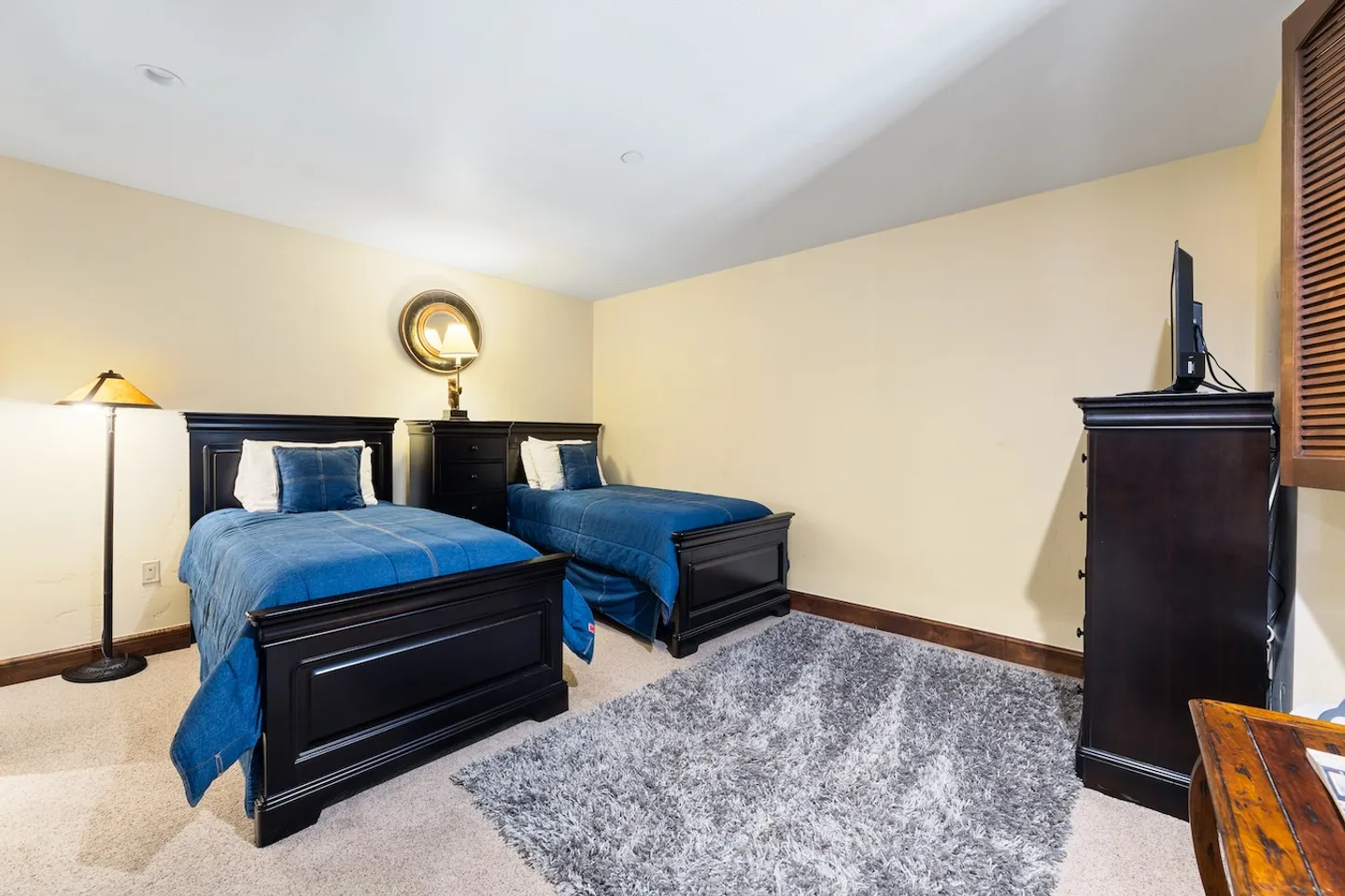 Bedroom with two twin beds featuring dark wood headboards and blue bedding, placed on light carpet with a gray shag rug.