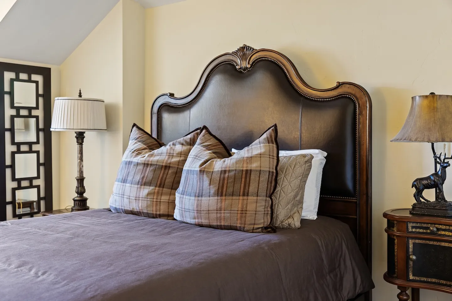 Close-up of a bed with a dark leather and carved wood headboard, plaid pillows, and adjacent nightstands with lamps.