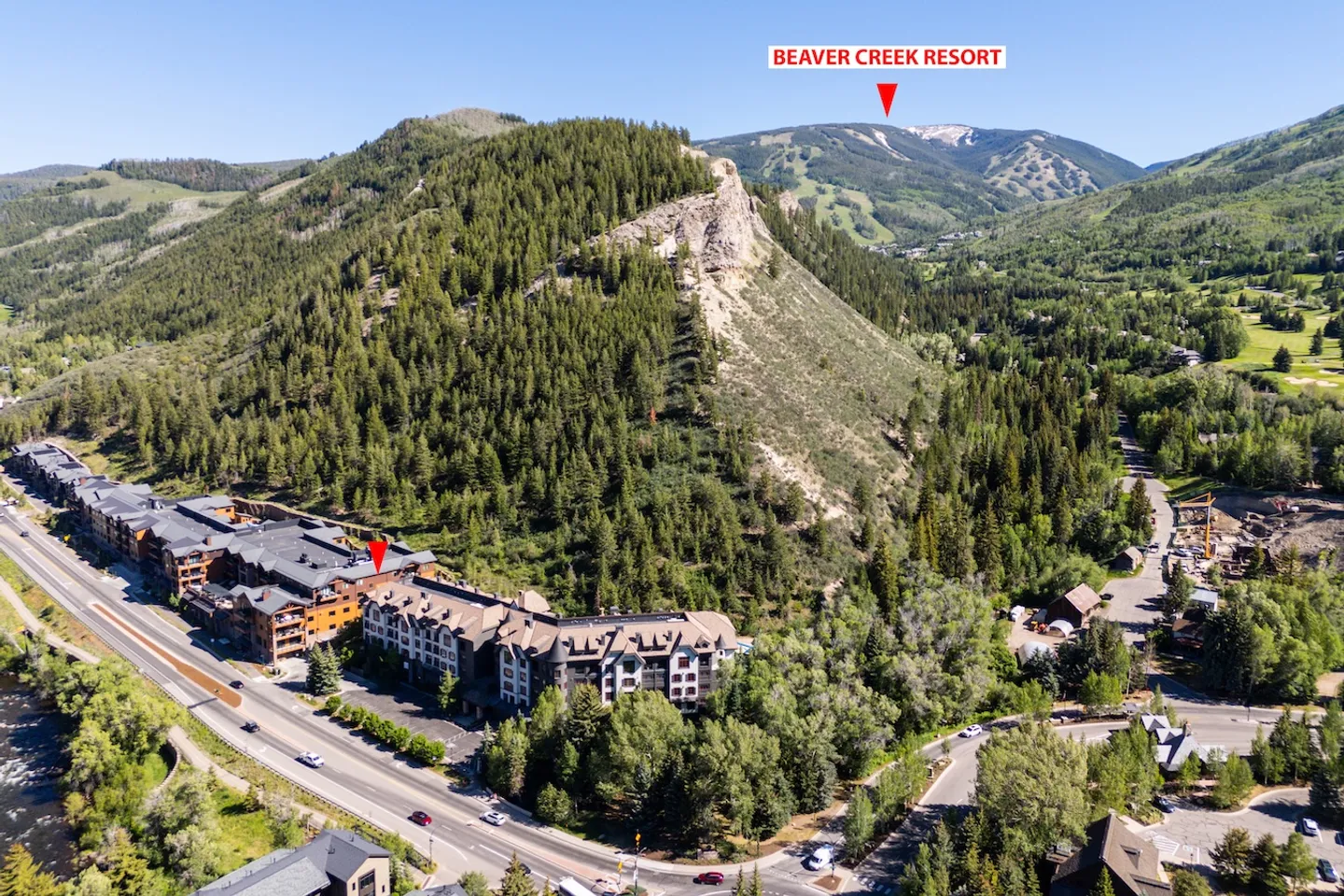 Aerial view of a multistory mountain lodge complex along a roadway, with forested hills and Beaver Creek Resort ski runs in the background under a blue sky.