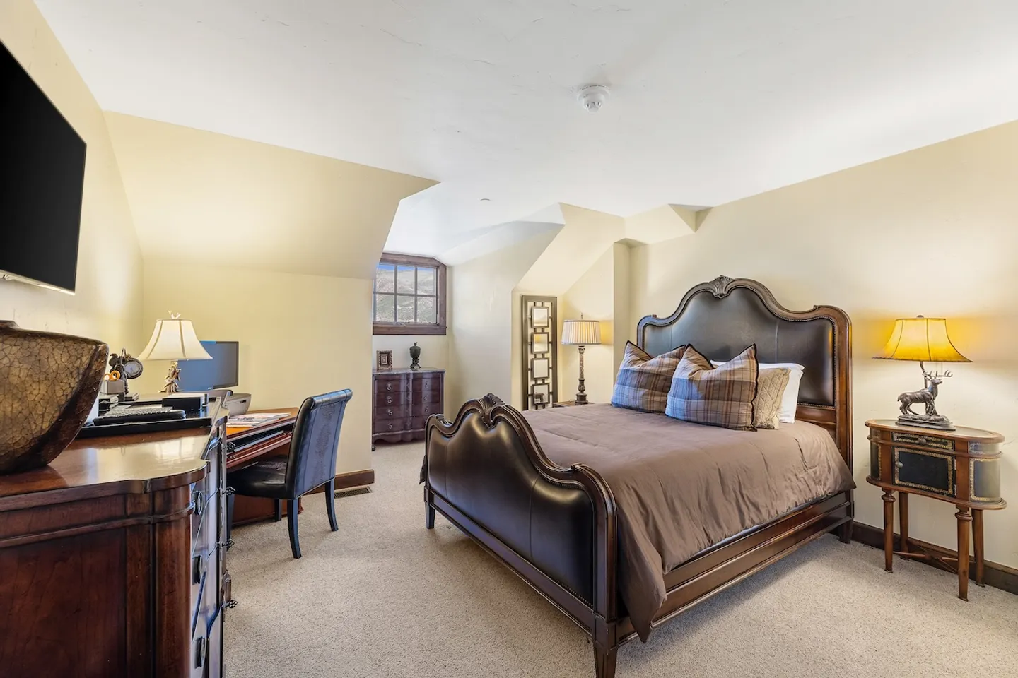 Bedroom with vaulted ceilings, a dark wood and leather bed, nightstands with lamps, a desk with a chair, and a dresser under a wall-mounted TV.