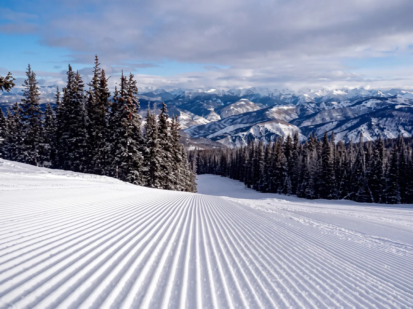 Freshly groomed corduroy snow on a ski slope flanked by evergreen trees with distant mountain ranges under a partly cloudy sky.