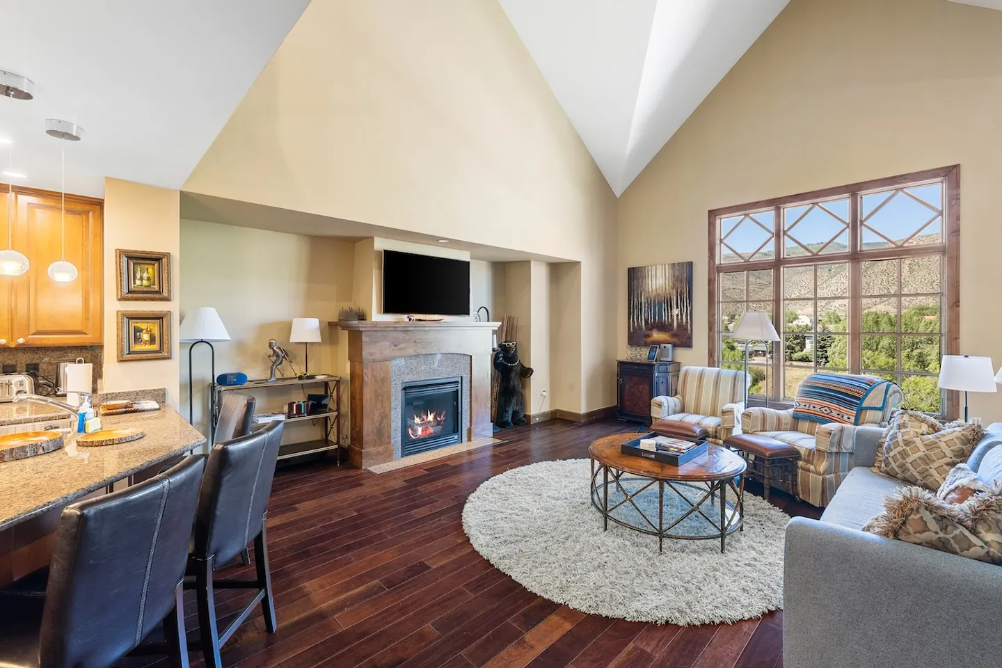 Open-concept living room with vaulted ceiling, wood floors, fireplace with TV above, and seating area next to a breakfast bar.
