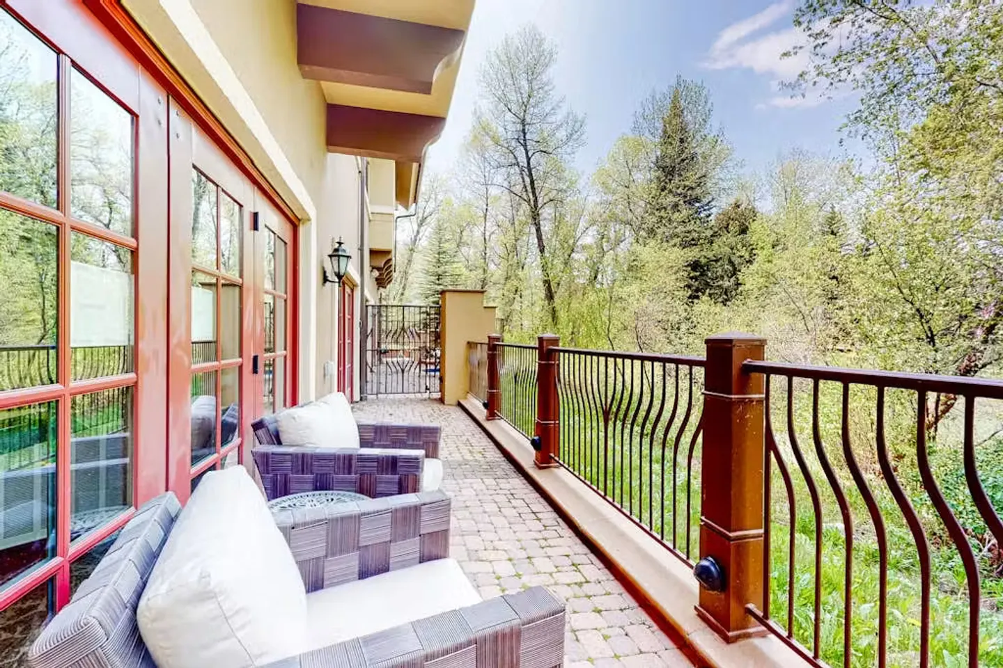 A long paved outdoor patio with woven armchairs, dark railing, and views of surrounding green trees.