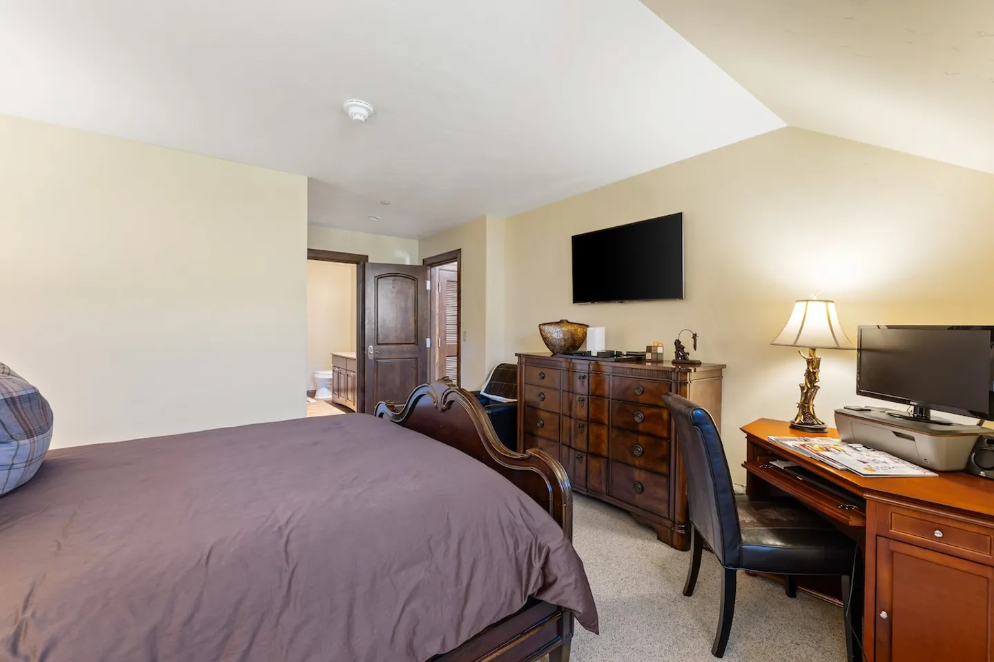 Bedroom with a wooden bed, dresser with a mounted television above it, and a study desk with a chair and monitor.