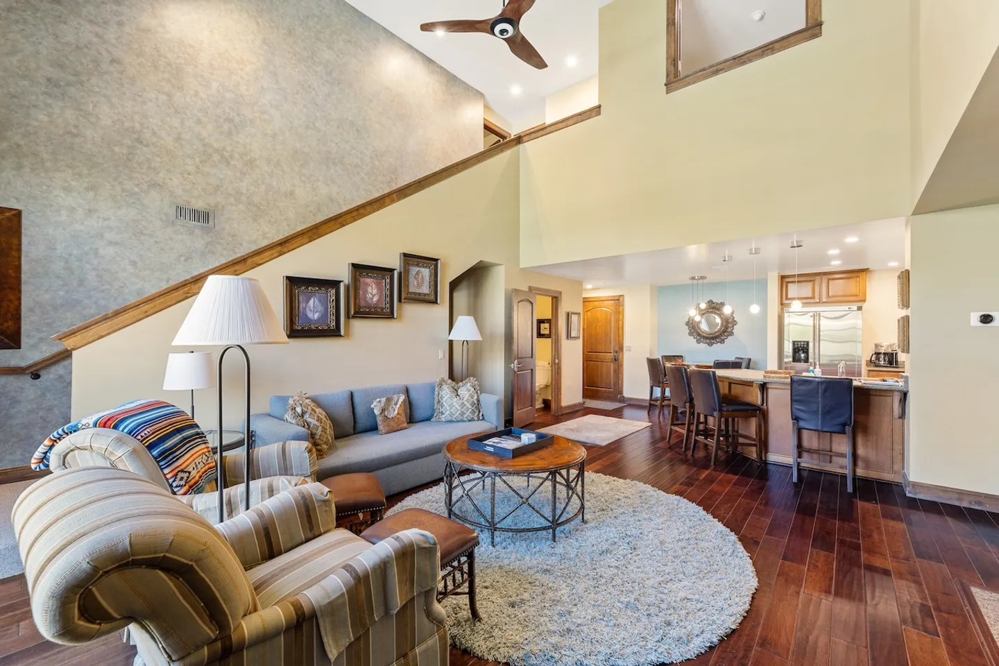 Open-concept living room with high ceilings, plush seating, round coffee table on a shag rug, and adjoining kitchen with a breakfast bar.