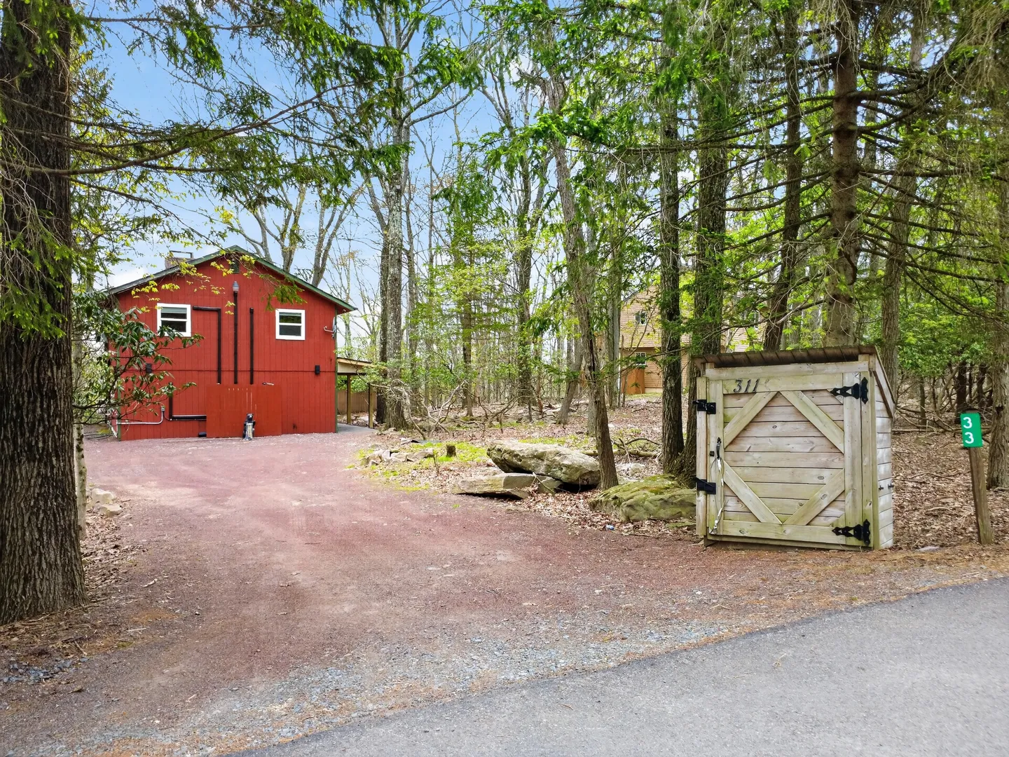 A gravel driveway leading past a wooden enclosure toward a two-story red building surrounded by trees.