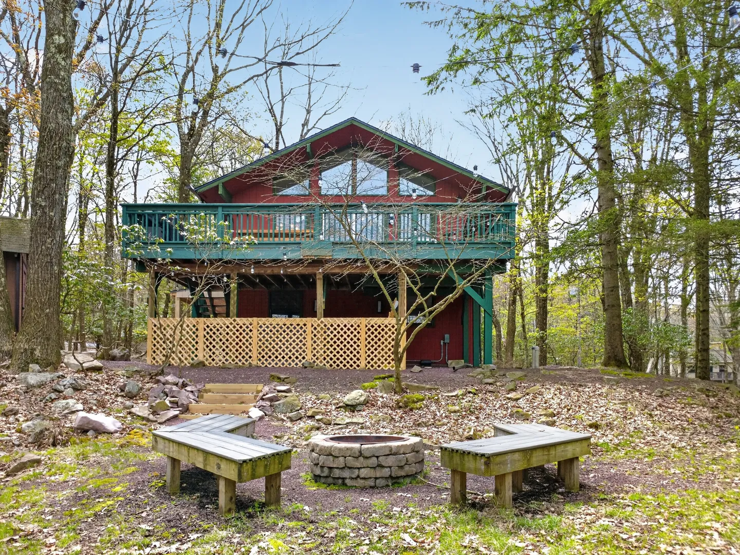 Exterior of a red two-story cabin surrounded by trees, featuring a large elevated green deck and a stone fire pit with wooden benches in the yard.