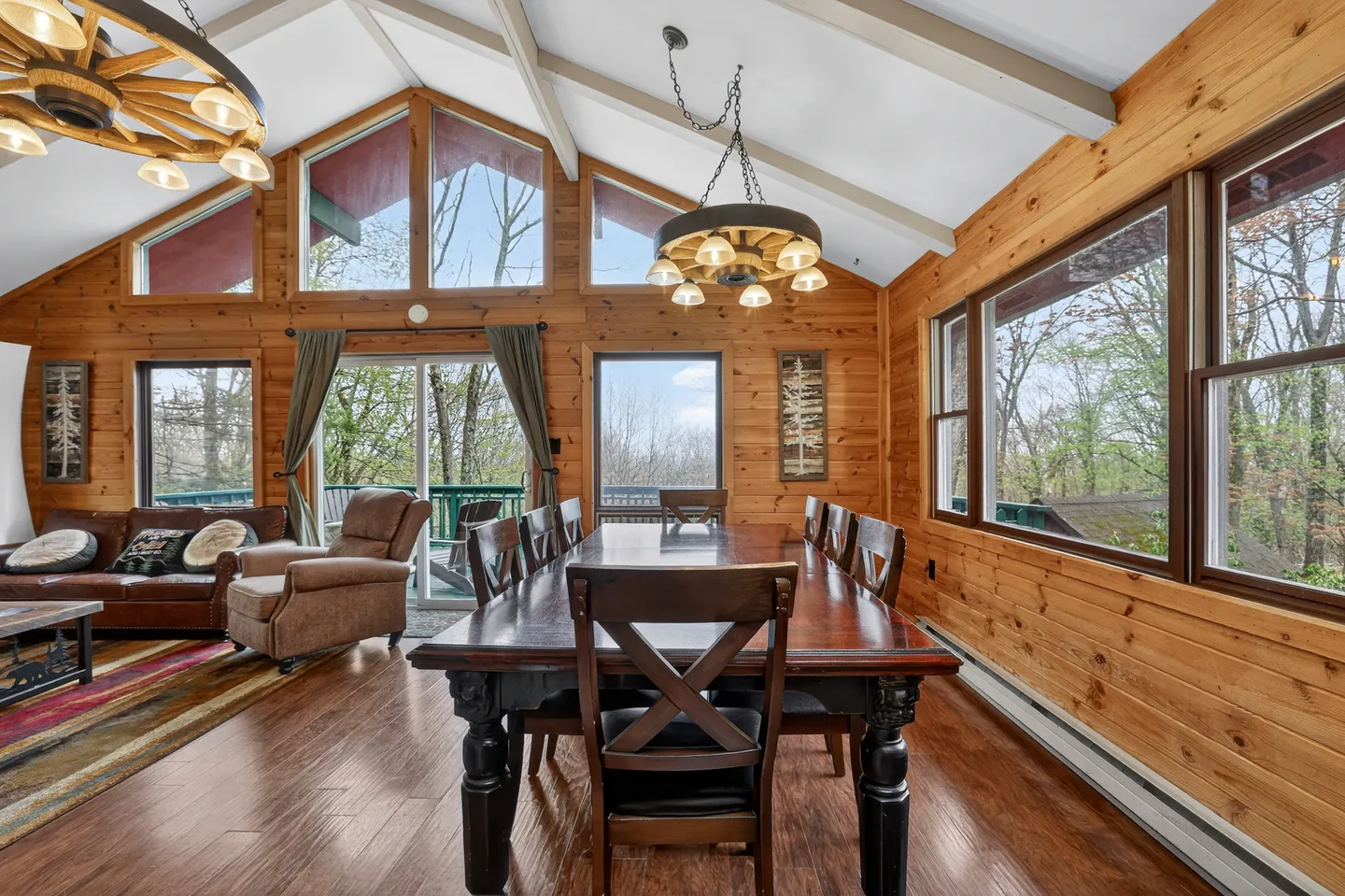 Dining area with a large wooden table, wagon-wheel chandeliers, wood-paneled walls, and adjacent living space with views to an outdoor deck.