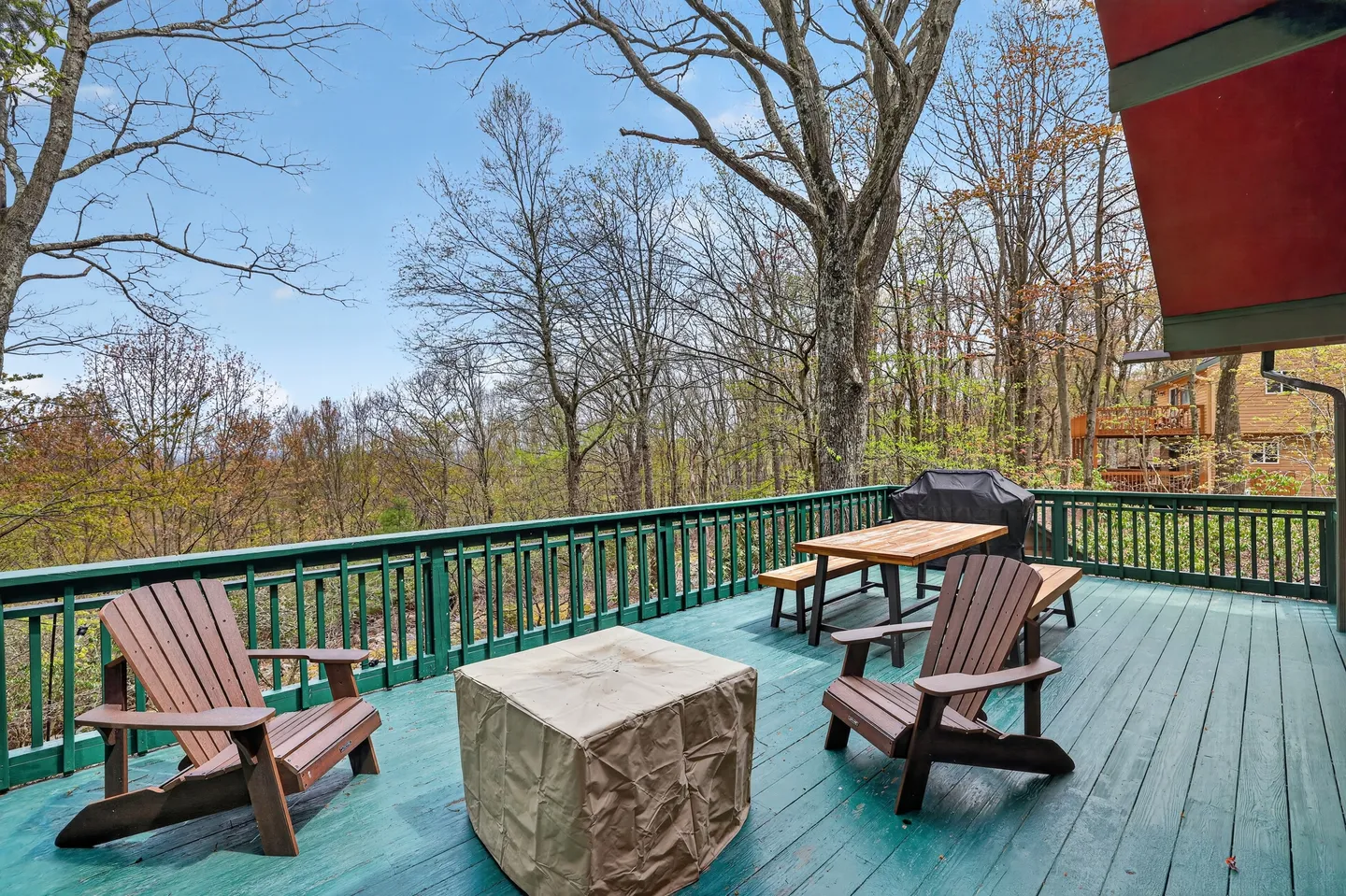 Outdoor wooden deck with wooden chairs, a covered fire pit or table, a picnic table, and a grill, surrounded by trees.
