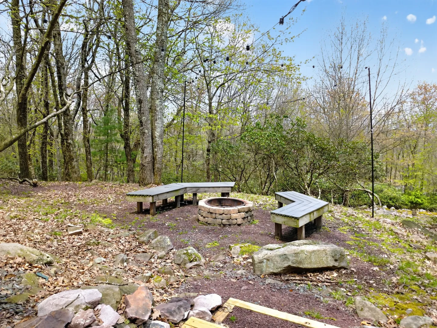 Outdoor stone fire pit surrounded by angled wooden benches beneath string lights in a wooded setting.
