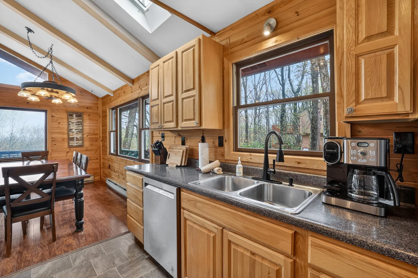 Kitchen counter with stainless steel sink, coffee maker, and dishwasher, leading into an open dining area with wood-paneled walls.