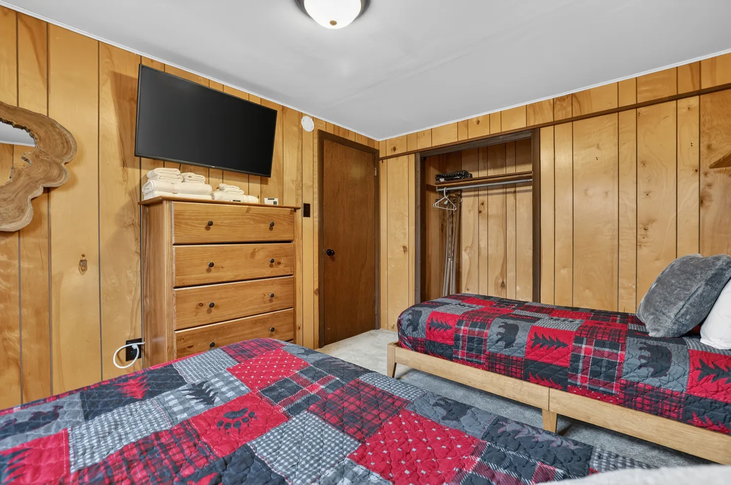 Bedroom with wood-paneled walls, twin beds with rustic red and black quilts, a wooden dresser with a mounted TV, and an open closet.