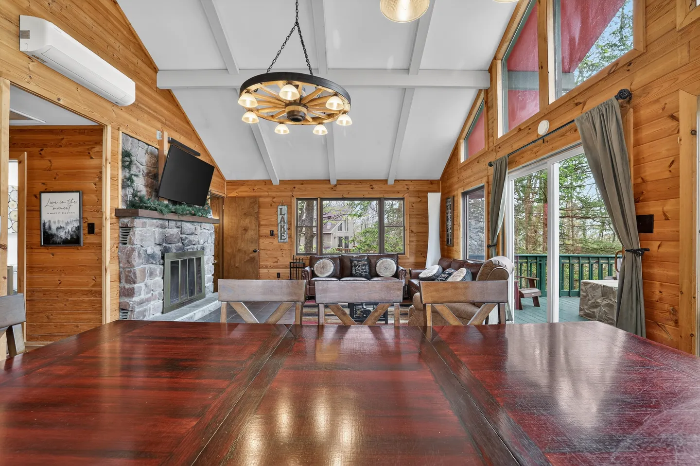 Open-concept dining and living room in a log cabin with a stone fireplace, wagon wheel chandelier, vaulted ceiling, and sliding glass doors leading to a deck.