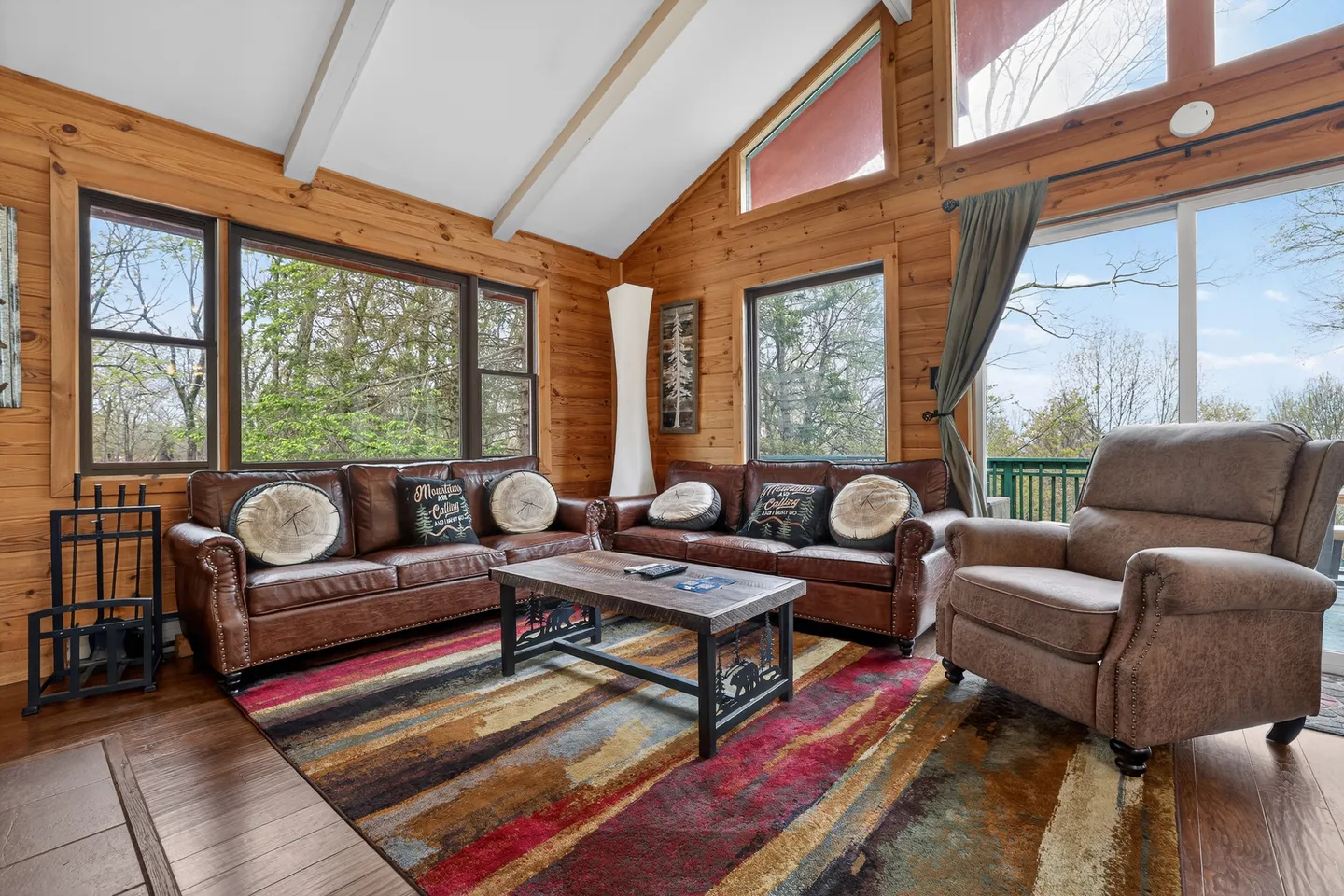 Living room with wood-paneled walls, vaulted ceiling, two brown leather sofas, an armchair, and large windows.