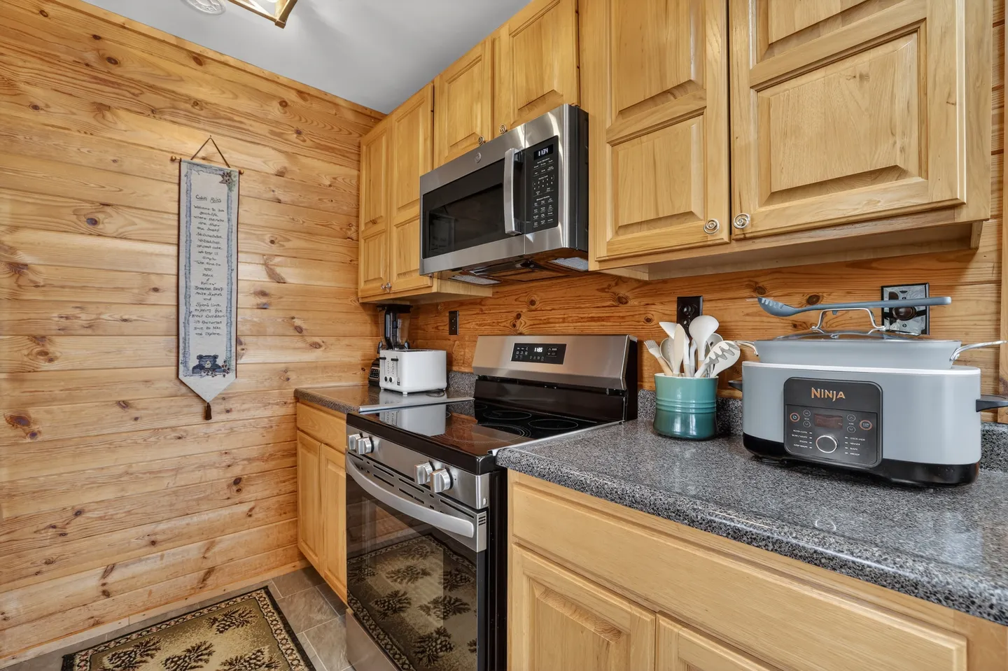 Kitchen area featuring wood paneled walls, light wood cabinetry, speckled countertops, a stainless steel range, and a microwave.
