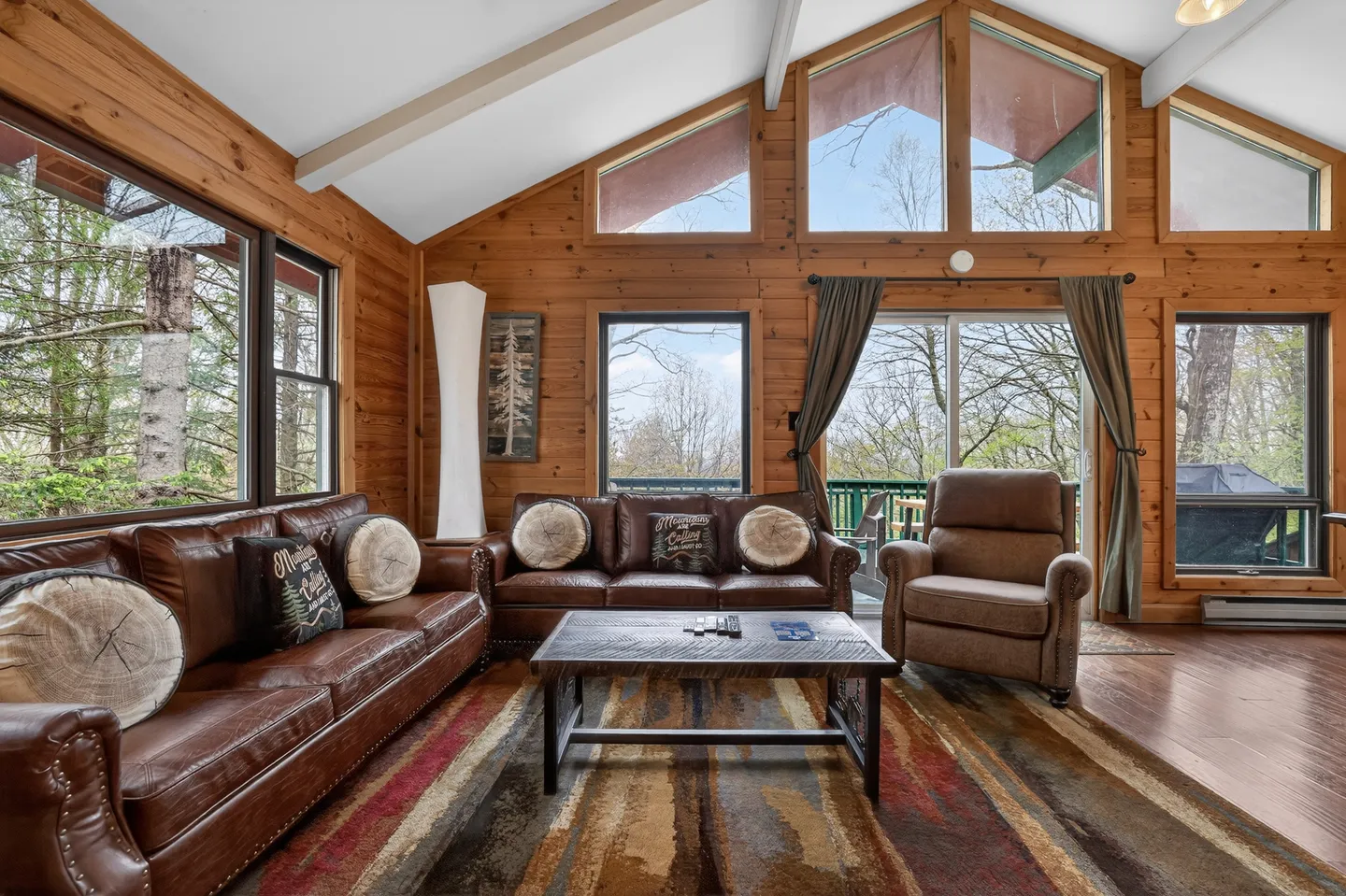 Living room with wood-paneled walls, vaulted ceiling, brown leather sofas, patterned rug, and large windows looking outside.