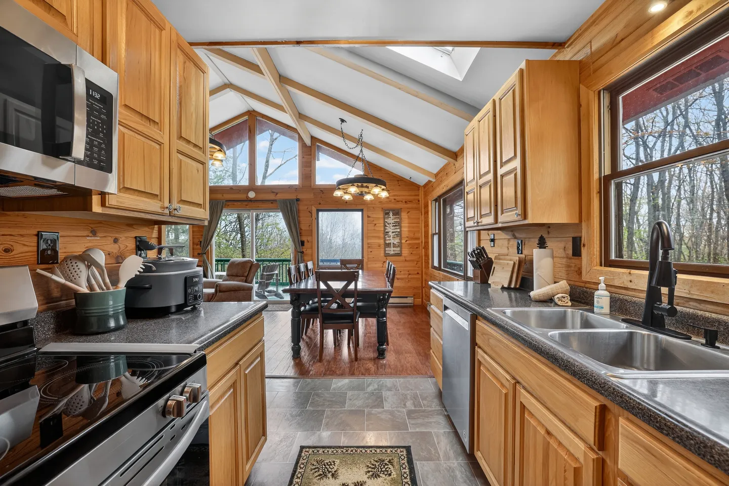 A kitchen with wooden cabinets and dark countertops looking into an open dining area with vaulted ceilings and large windows.