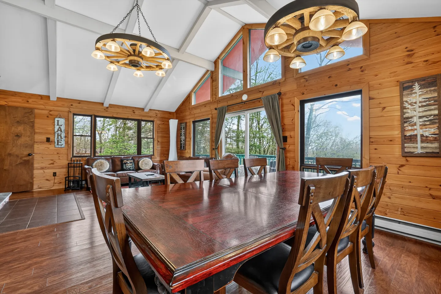 Dining area with a large wooden table, wagon-wheel chandeliers, wood-paneled walls, and views of the living space and deck.