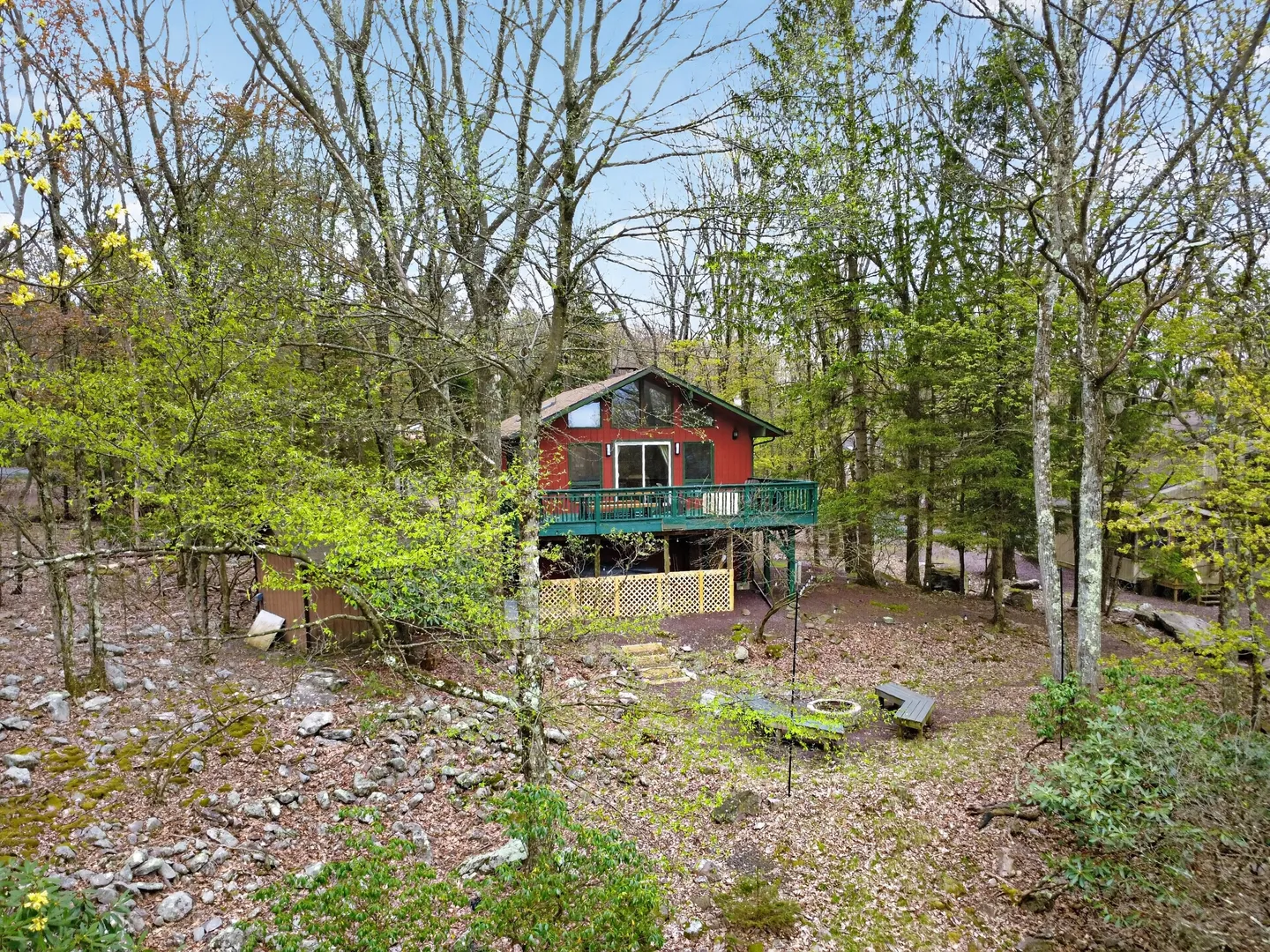 Exterior view of a red two-story cabin with a green elevated deck, surrounded by dense trees, rocky ground, and a fire pit area.