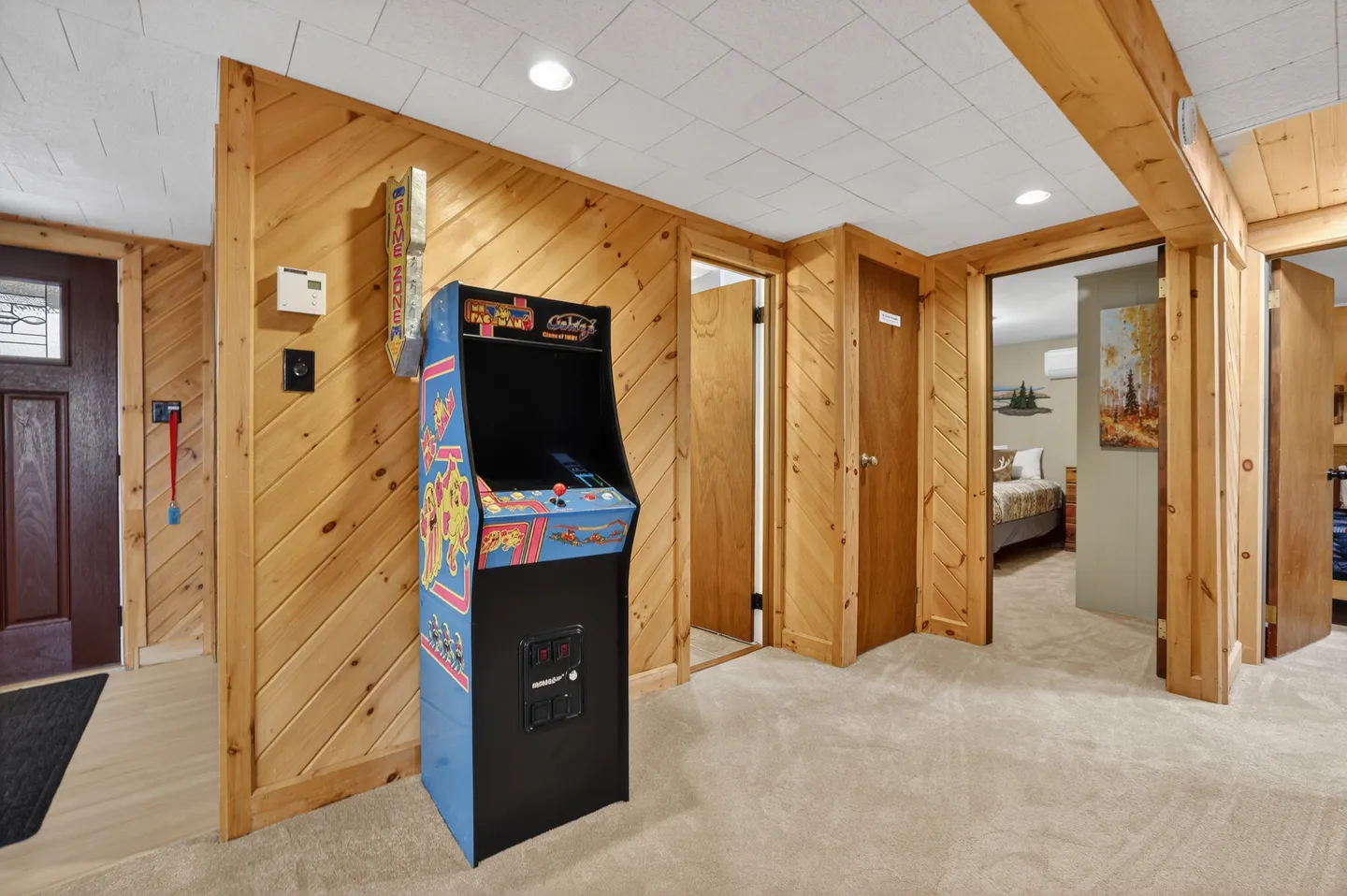 Hallway featuring knotty pine wood-paneled walls, carpet flooring, a standalone arcade machine, and doorways leading to adjacent rooms.