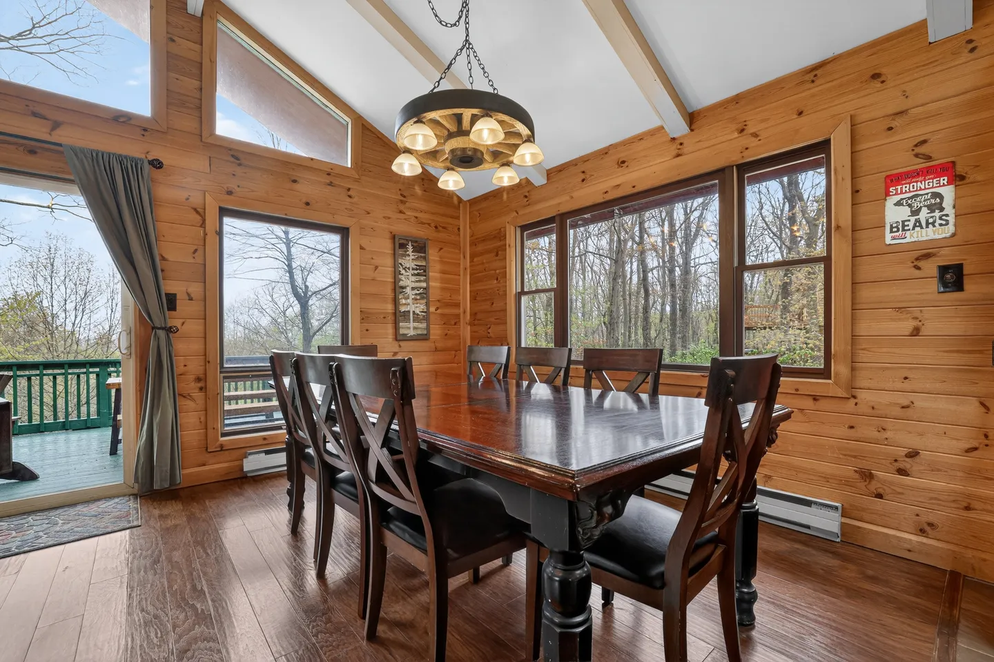 Dining area with wood paneled walls, a large wooden table with chairs, a wagon-wheel style light fixture, and windows looking out into the trees.