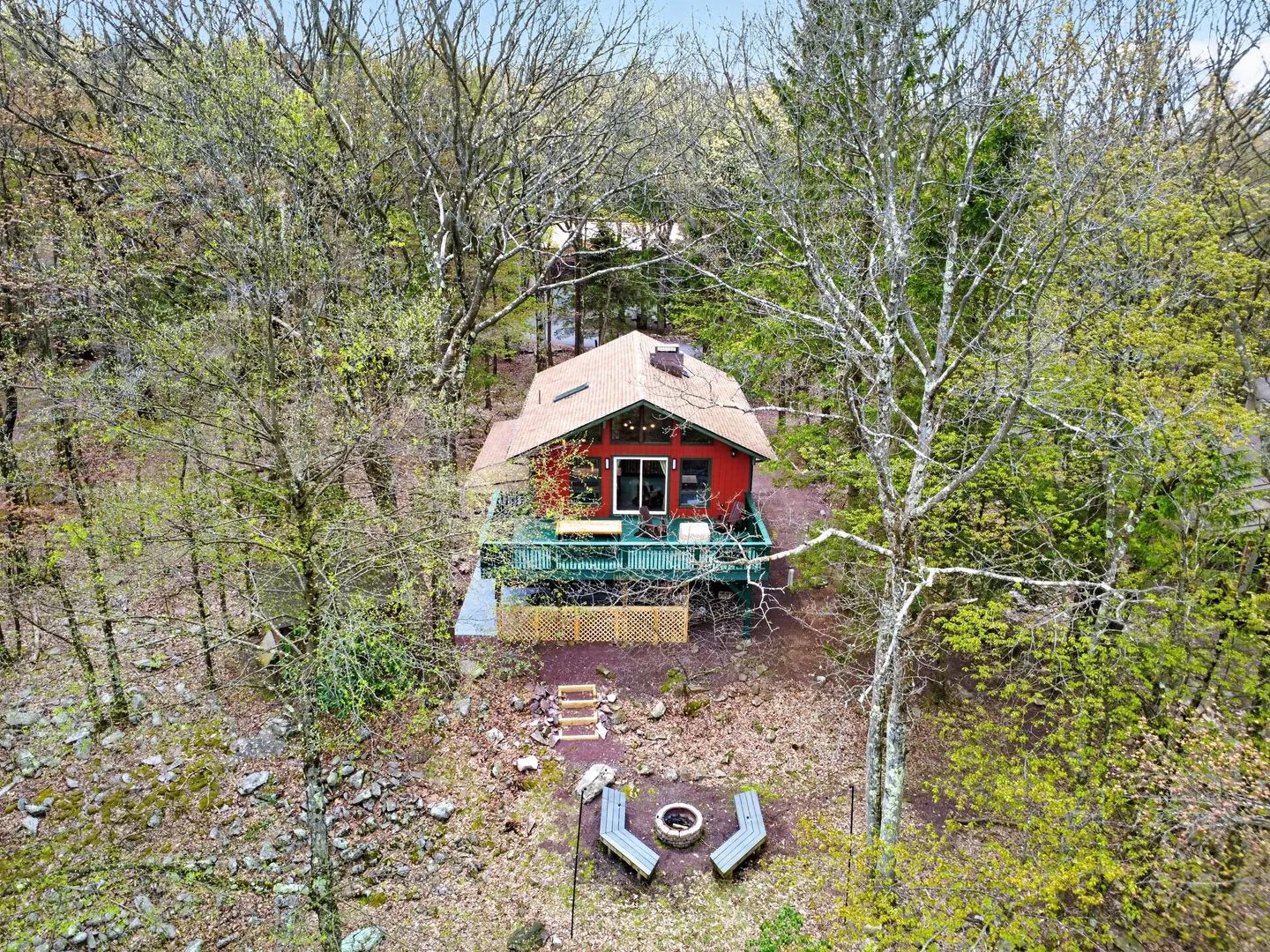 Elevated view of a red cabin in the woods with a raised deck and a fire pit area with curved benches below.