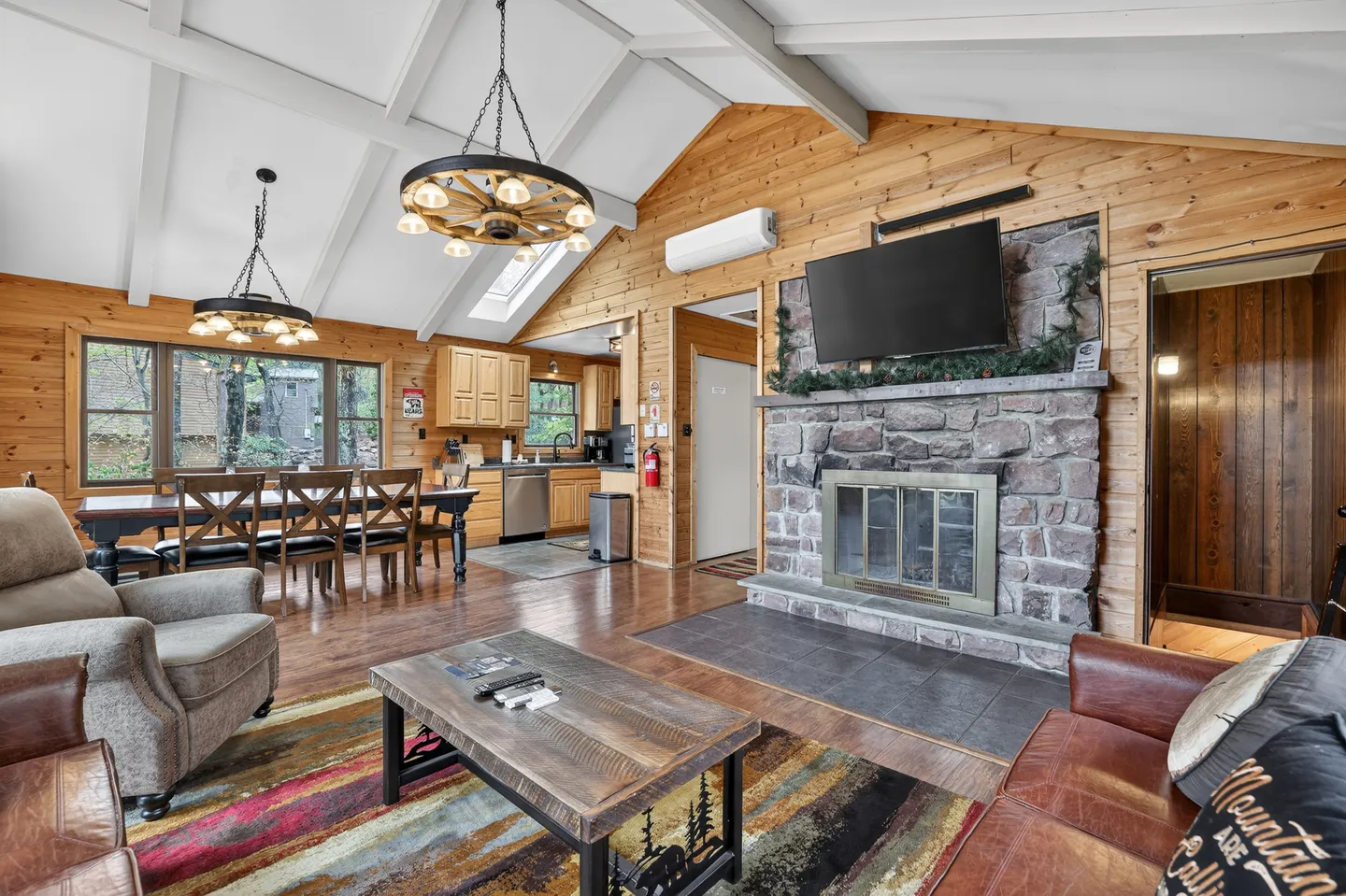 Open living area with a stone fireplace, wood-paneled walls, vaulted ceiling, and view into the dining space and kitchen.